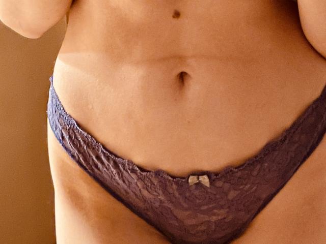 SweetClaraa's flat, tan-lined belly with a small navel, wearing purple lace panties with a tiny white bow, showing her soft, slightly curvy, lower abdomen.