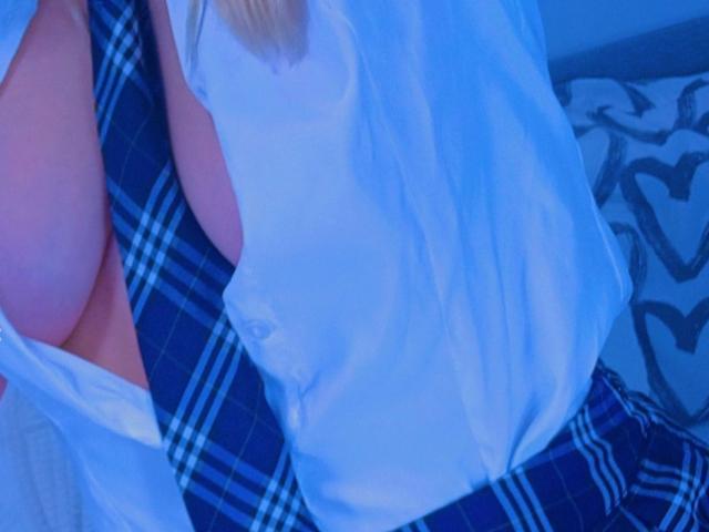 La camicia bianca di LiluL è aperta, rivelando un seno generoso, e indossa una gonna a quadri blu. I suoi capelli biondi sono parzialmente visibili. L'immagine ha una tonalità blu..