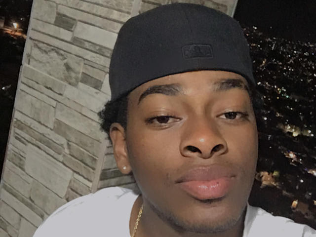 JackConnor, un uomo nero con una carnagione media, indossa un berretto nero da baseball, una maglietta bianca e una catena d'oro, e sembra leggermente annoiato, con un sorriso appena accennato..