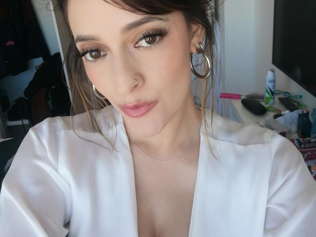 JaneRoa with brown eyes, nose ring, and pink lips, wearing a white shirt, silver necklace, and hoop earrings, has a subtle smirk and a slight chin lift.