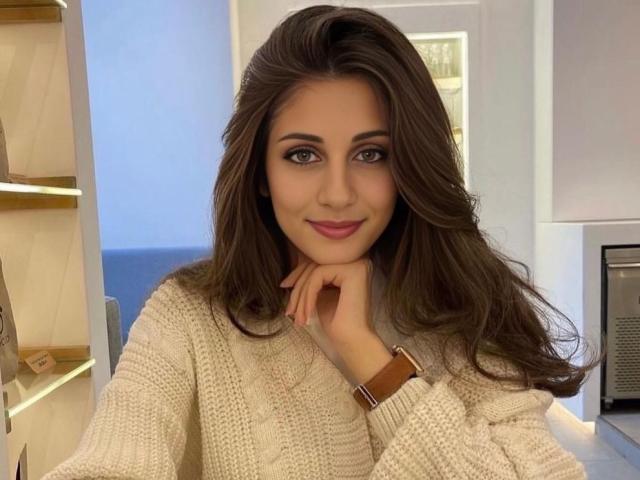 MarinaSexyAndHot with long brown hair, wearing a cream knit sweater, brown watch, and pink lipstick, smiling with hand on chin, looking directly at camera.