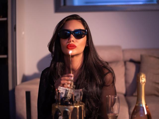 AlessandraMalory with long black hair, red lips, and dark sunglasses, blowing a match while holding a champagne bottle, a glass, and a golden bucket.