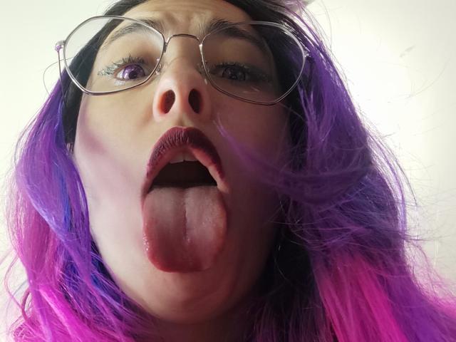 NatashaLau with purple and pink hair, glasses, and tongue out, looking up at the camera. Her mouth is open, showing her tongue and red lips.