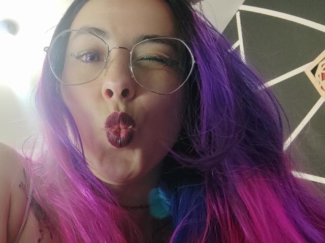 NatashaLau with purple and pink hair, wearing glasses, puckering her lips in a kiss, showing her plump lips and slightly winking with green and purple eye makeup.