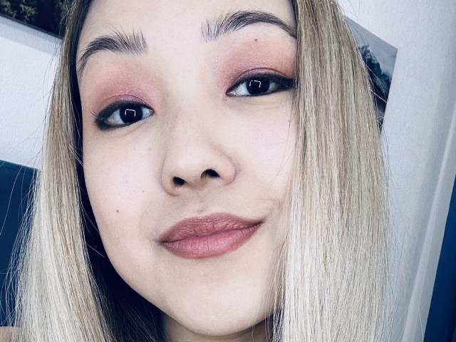 RenyLime's face with blonde hair, pink eyeshadow, black eyeliner, and pink lips, smiling slightly, showing a cute and sexy look with her small nose and smooth skin.