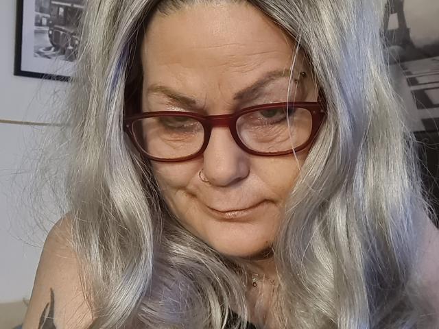 LatexShantie with gray, wavy hair, red glasses, nose ring, and slight smile, showing tattoo on right shoulder, looking down. Boobless, no visible ass or pussy.