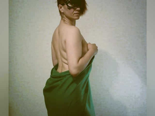 LoriFixS with messy brown hair, wearing a black mask, stands topless, covering ass with a green towel, showing a slender back and a slightly turned, serious face.