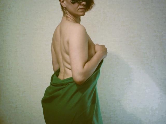 LoriFixS is topless, covering her ass with a green towel, back facing the camera, wearing dark sunglasses, with short brown hair and a slightly grim face.