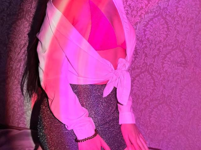 Los pechos de LisaAlisia en un sujetador rosa, una camisa blanca atada al frente y una falda gris brillante, con cabello negro largo, iluminación rosa y morada, y una pulsera de plata..