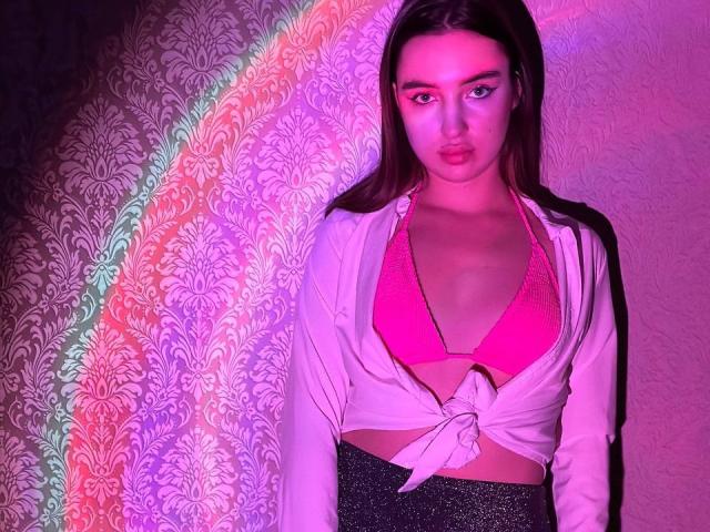 LisaAlisia con un sujetador rosa, una camisa blanca atada debajo de los senos, pantalones negros brillantes, iluminación rosa y morada, cabello castaño largo, expresión de labios fruncidos, de pie, cuerpo completo, sexy, segura de sí misma..