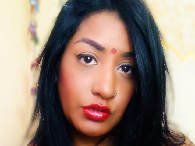 Nahirr has dark black hair, red lipstick, and a small red dot on her forehead. She's wearing a nose ring and has thick, arched eyebrows. Her makeup is bold.
