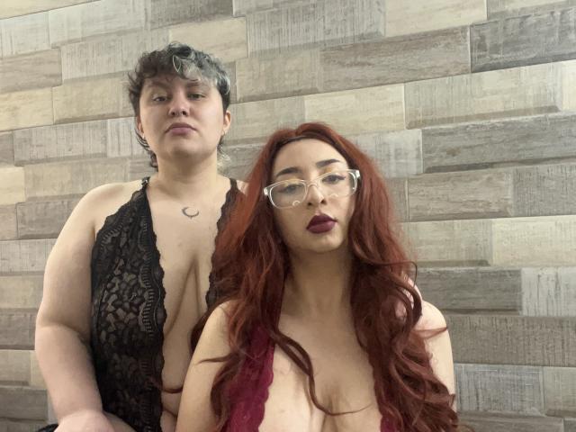 RavenAndEllie with short dark hair and black lace lingerie standing behind RavenAndEllie with long red hair, glasses, and red lace top, showing big tits and small ass.