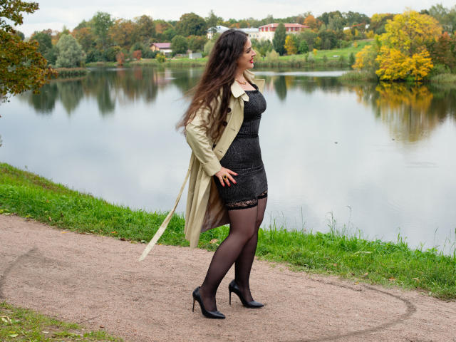 MimmyLongNipples stands on a gravel path, wearing a green coat, black lace dress, black stockings, and high heels. She has long brown hair, a curvy ass, and black heels.