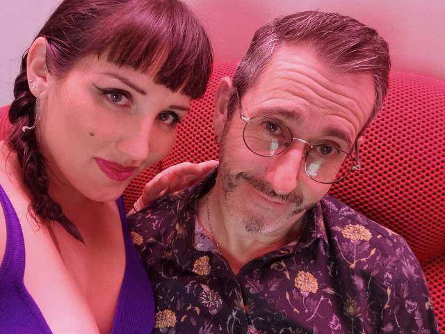 TuttiFloyd69 with big boobs in a purple top and TuttiFloyd69 with glasses, mustache, and floral shirt, sitting on a red chair, both smiling at the camera.