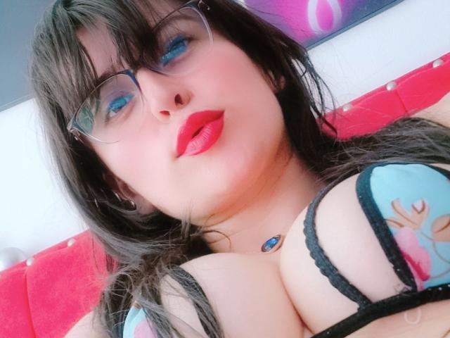 LindaSmith with blue eyeshadow, red lipstick, glasses, and black lace bra, showing her big tits, wearing a blue gem necklace, lying on a red surface.