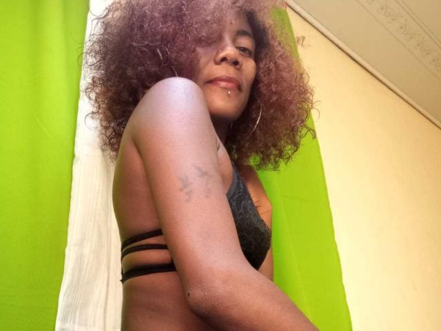 GinaSensual69 con capelli ricci castano scuro, indossa un reggiseno di pizzo grigio, mostra un livido sul braccio destro, ha un piccolo piercing al naso e uno sfondo con una tenda verde..