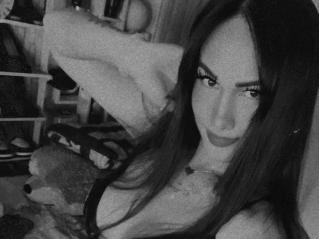 TsNaughtMiss69 with long dark hair, looking seductively at the camera, wearing a low-cut top showing her tits, in a black and white selfie, arm raised behind her head.