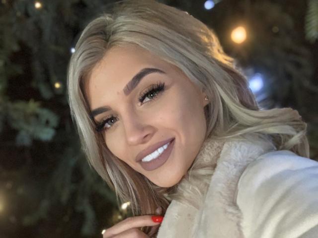 HottieLore has blonde hair, full eyebrows, long lashes, and is smiling with white teeth. She's wearing a white fur coat, and her red nails are visible.