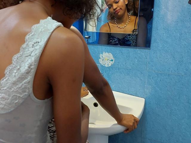 LouisaRiynah with curly hair, wearing a white lace tank top, washing her hands in a blue-tiled bathroom, reflected in the mirror, showing her dark-skinned, busty, pearl-necked chest.