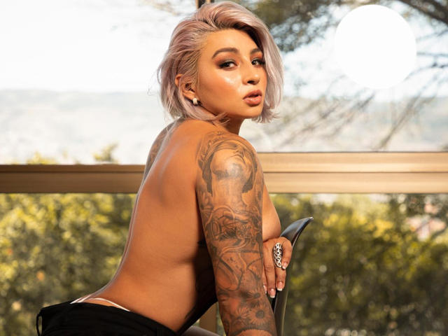 AvrilGray with pink hair, topless, showing a tattooed arm, looking back, with a pouty face, wearing black bottoms, holding a chair, outside with a blurred forest background.