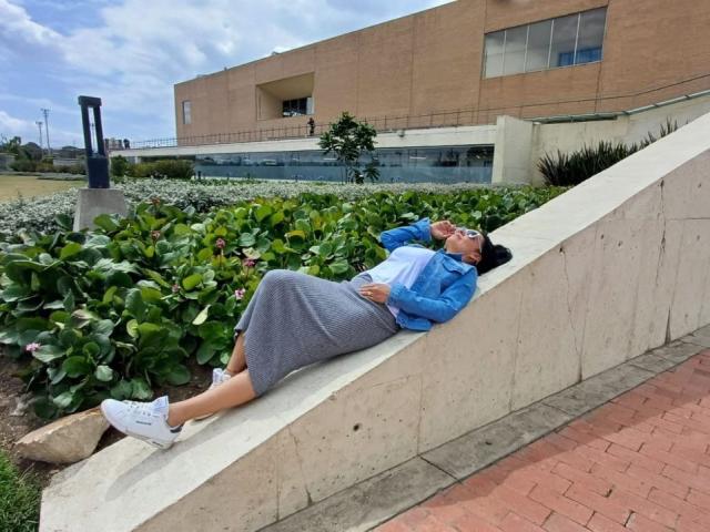 LailaCastillo is chillin' on a concrete slope, rockin' a blue jacket, white shirt, gray skirt, and white sneakers, with her ass up and legs down, talkin' on phone.