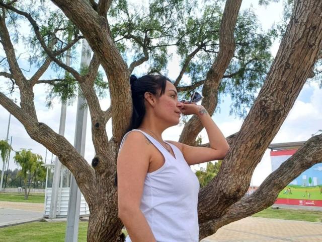 Laila Castillo, met zwart haar in een staart, een witte tanktop, die uit een flesje drinkt, staand tegen een grote boom, rechterzijprofiel, zonnige dag, blauwe lucht..