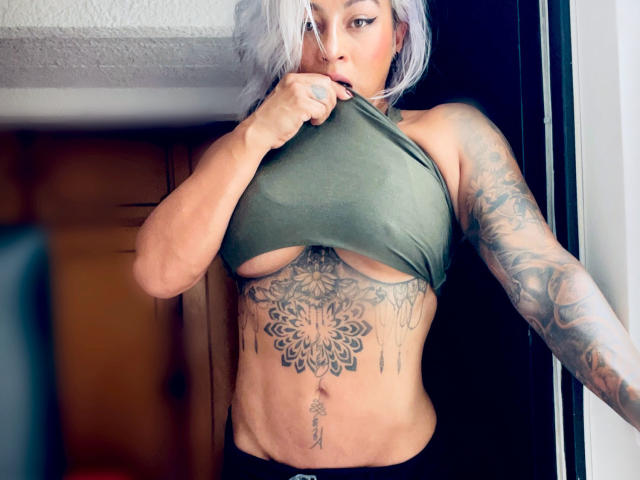 AvrilGray with silver hair, lifting a green top to expose her tattoos, including a large mandala on her chest and arms, showing her tits and toned abs.