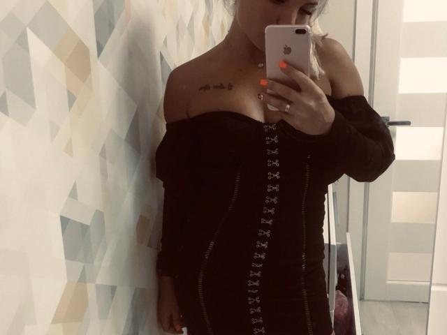JudyGrace with blonde hair, off-shoulder black dress, lace-up front, holding iPhone, showing small tattoo above boob, nails painted orange, mirror selfie, room with patterned wall.