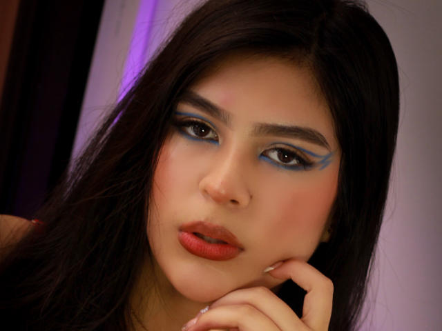 SamanthaEva69 has dark brown hair, blue eyeshadow, and red lips, with a pensive expression, resting her chin on her hand. Her pink cheeks and brown eyes are visible.