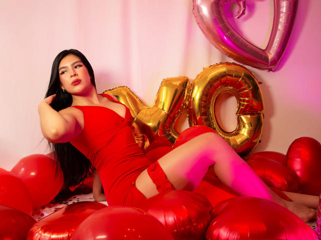 SamanthaEva69 with long black hair, red dress, and garter, sitting on red balloons, surrounded by gold "NO" balloons, red heart balloons, and pink lighting. Her ass is prominent.