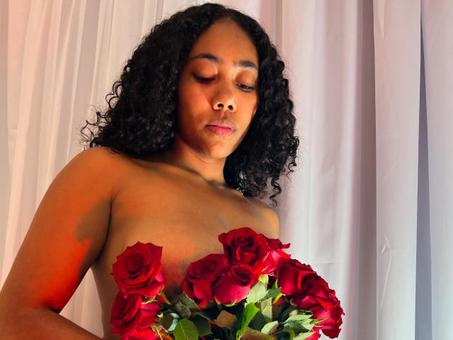 AishaRhodes with curly black hair, naked, holding a bouquet of red roses covering her tits, looking down with closed eyes, soft orange light on her right side.