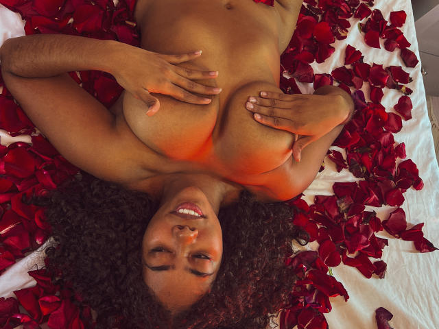 AishaRhodes, nude with curly black hair, lying on red rose petals, covering her big tits with her hands, smiling, dark brown skin, and red light on her body.