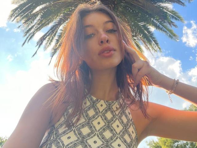 Arikay con lunghi capelli castani, indossa una canotta senza maniche con motivo a rombi, con i capelli illuminati dal sole, una mano vicino al viso, cielo blu, nuvole bianche, palma, sorriso dolce, leggero alone luminoso..