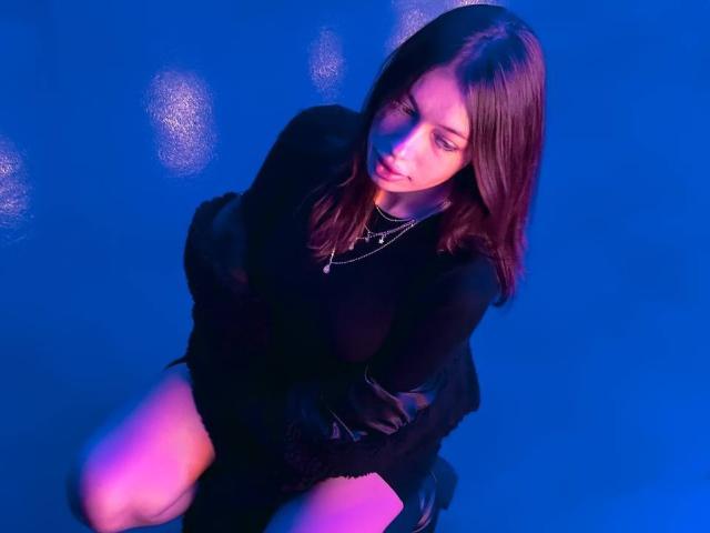 Arikay, com cabelo castanho médio, vestido preto e colar, ajoelhada com os olhos fechados, mostrando o bumbum, sob iluminação azul e rosa..