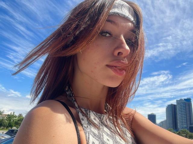 Arikay, con i capelli castani, una fascia bianca in testa e una maglietta a fantasia, mostra il suo viso e le labbra piene. Sullo sfondo, un cielo azzurro e gli edifici della città. Il suo seno è piccolo..