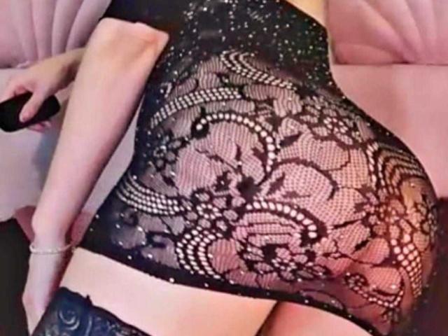 BlueNightX wearing a black lace top that shows her tits, with intricate floral patterns and a slightly open bottom, holding a black object in her right hand.