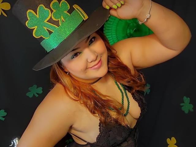 RouseWatsons with orange hair, wearing a glittery green hat with three clovers, black lace bra, green necklace, and green nails, smiling at the camera, showing her tits.