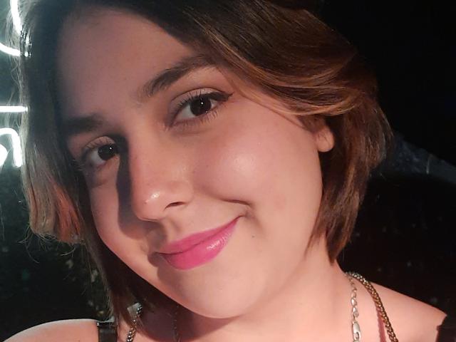 AlessaMih's face, showing a cute smile with pink lipstick, short brown hair, and a shiny nose. She's wearing a necklace and has a soft, fair skin tone.