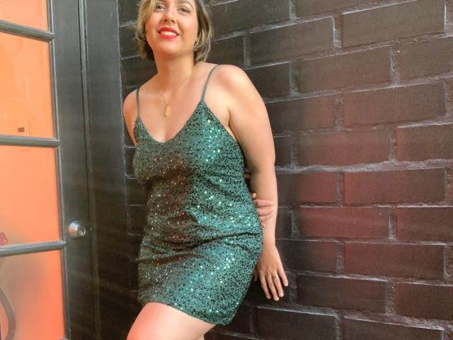 AlessaMih, curvy with medium tits, wearing a green sequined dress, leaning against a dark brick wall, smiling with red lipstick, short blonde hair, and a gold necklace.