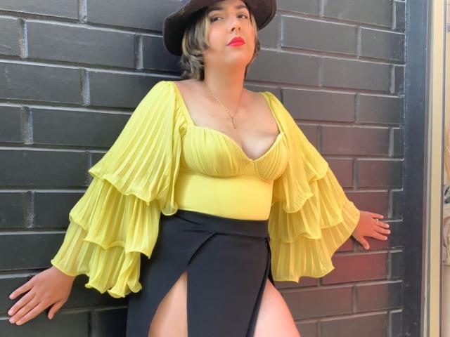 AlessaMih with yellow ruffled top, black high-waisted skirt with thigh slit, black hat, red lipstick, and short blonde hair, leaning against black brick wall. Boobs visible.