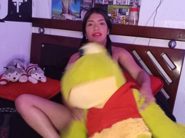 ArianithaXox with medium tits, dark hair, and red lipstick, sitting on a bed, holding a yellow and red stuffed frog, with stuffed dogs to her left.