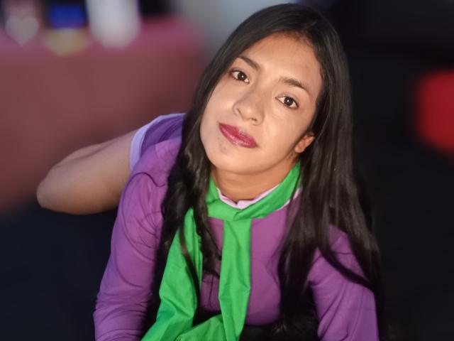 ArianithaXox with long black hair, purple shirt, green scarf, and pink lipstick, looking up with a slight smile, showing her brown eyes and subtle makeup.