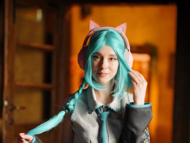 MissAstravert with teal hair, cat ears, pink headphones, holding braid, smiling, wearing white shirt with teal tie, in warm light, holding hair with right hand, looking cute.
