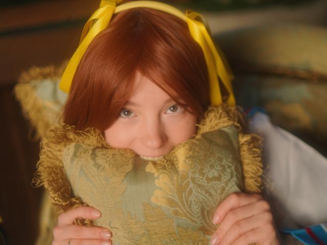 MissAstravert with red hair and yellow headband, holding a gold, fringed pillow, her cute face peeking above, looking at the camera with a gentle smile.