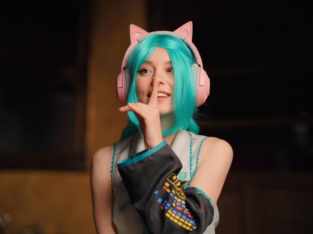 MissAstravert with teal blue hair, pink cat ears, and pink headphones, touching her lips, wearing a grey and black cosplay outfit, with a cute and playful expression.