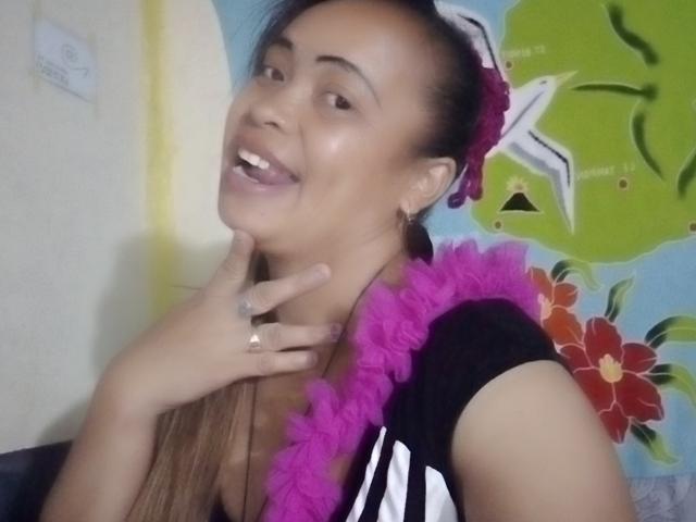 Rahel69 con cabello castaño largo, un collar de flores rosa, una camisa negra y blanca, sonriendo, tocándose el cuello, mostrando un anillo y llevando una flor rosa en el cabello..