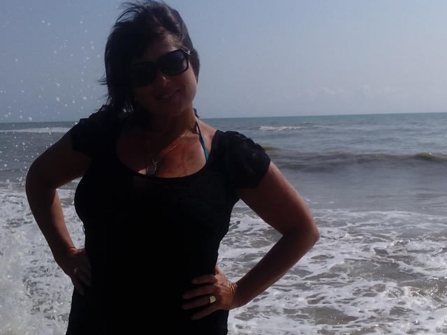 Ellita stands at the beach, wearing a black dress, sunglasses, and a necklace, with hands on her hips, showing her tits and smiling, water splashing around her.