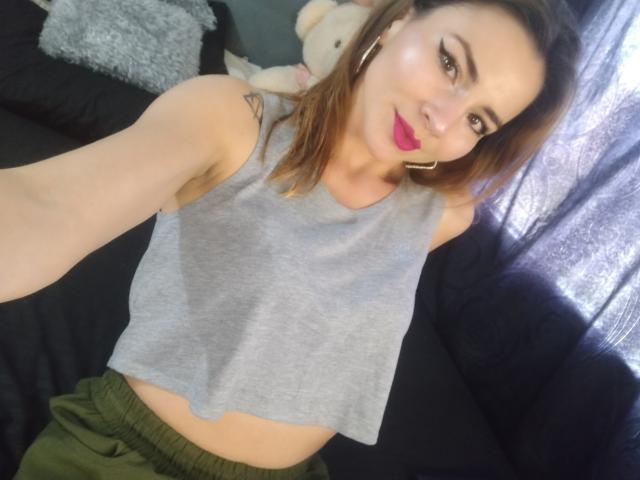 EmelySofia with pink lipstick, gray sleeveless top, small tits, green pants, brown hair, gold hoop earrings, tattoo on right shoulder, smiling, black couch, stuffed bear in background.