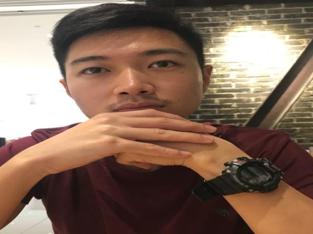 ChristopherLongest with short black hair, a small mustache, and a dark maroon shirt, biting his right hand, wearing a black wristwatch, looking at the camera.