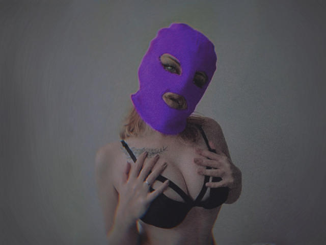 MissNestie with purple mask, black bra, and hands on chest, showing her tits and tattoo. She's got blonde hair, and a sexy look, standing in a gray room.